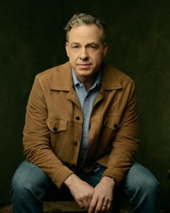 Jake Tapper sits wearing blue jeans a light blue button down shirt and a tan jacket